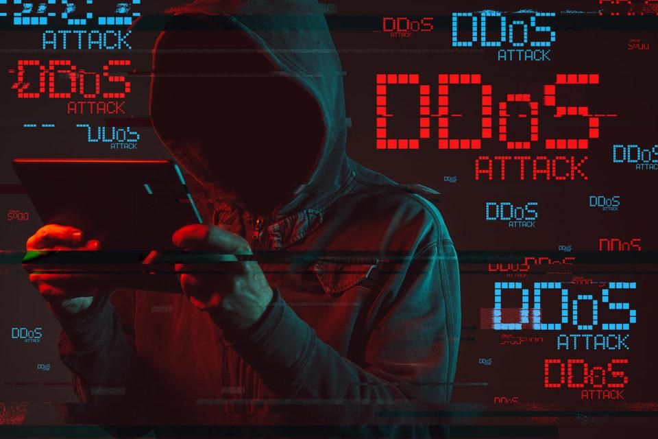 Silueta encapuchada frente a múltiples alertas de “DDoS Attack”, representando un ataque de denegación de servicio contra sistemas digitales.