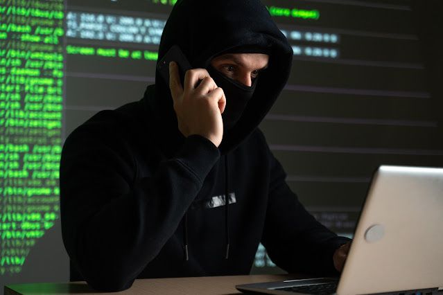 Persona encapuchada utilizando un ordenador portátil y un teléfono frente a pantallas con código, representando técnicas de ingeniería social y ciberdelincuencia.